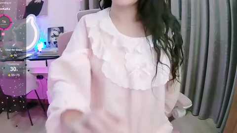 Media: Video of an Asian woman with long, wavy black hair, wearing a white, ruffled blouse, standing in a room with a desk, chair, and grey curtains in the background.