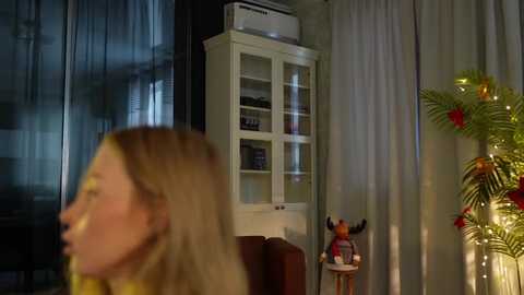 Media: Video of a dimly lit living room with a blonde woman in motion on the left, a white cabinet with glass doors, a red chair, and a decorative plant on the right.