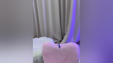 Media: Video of a plush heart-shaped pillow with a blue tag on the back, set against a backdrop of soft, flowing curtains in neutral tones with purple lighting. The scene exudes a cozy, intimate atmosphere.