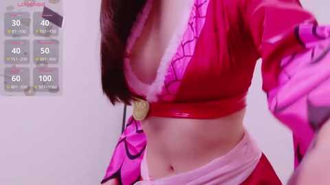 A video of a woman with long, straight brown hair, wearing a revealing, pink, spider-themed crop top with a gold clasp, and a matching skirt with black accents.