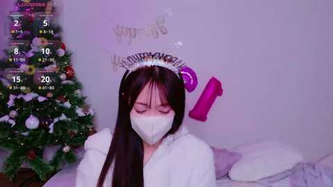 A video of an Asian woman with long black hair, wearing a white mask, a silver tiara, and a festive white hoodie. She's indoors, in front of a decorated Christmas tree.