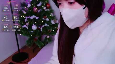 A video of a young woman with long dark hair and a white mask, wearing a fluffy white robe, sitting near a decorated Christmas tree in a dimly lit room.