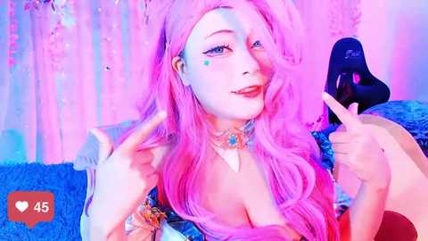 Media: Video of a fair-skinned woman with long, wavy pink hair and makeup, including blue and green eyeshadow and a heart-shaped blue eyeshadow on her cheek. She wears a black top, a choker with a heart pendant, and a blue beanie. The background is a pink and blue gradient with confetti.