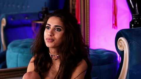 Media: Video of a young woman with long, wavy dark hair, wearing a brown top, seated in a teal velvet chair, in a dimly lit room with purple lighting.