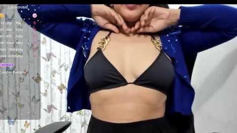 Media: Video of a woman with medium skin tone, wearing a black bikini top with gold chains and a blue cardigan. She has medium-sized breasts, and her hands are clasped behind her head. Background features a white curtain with a floral pattern.