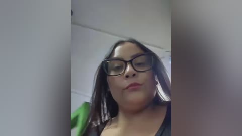 Media: Video of a light-skinned woman with long, dark hair and large, black-framed glasses, wearing a black top. She has a neutral expression and is standing indoors against a white wall, with a green garment hanging in the background.