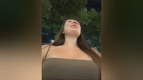 Media: Video of a young woman with long brown hair, wearing a strapless olive-green top, looking up, surrounded by lush green trees.