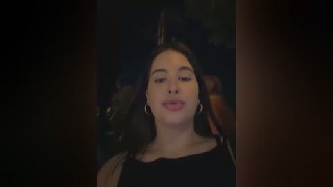 Media: A video of a young woman with fair skin, long dark hair, and subtle makeup, wearing a black top, against a dimly lit, blurred background.