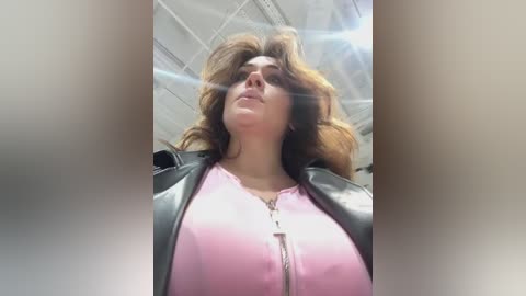 Media: Video of a woman with voluminous, wavy brown hair, wearing a pink top and black leather jacket, standing in a brightly lit room with a high ceiling.