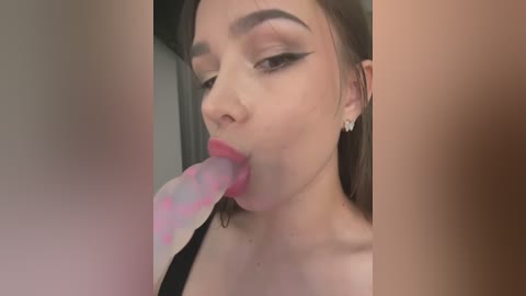 Media: Video of a young, light-skinned woman with dark hair and heavy makeup, licking a pink, polka-dotted lollipop, wearing a black top.