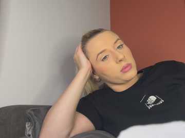 Media: Video of a blonde woman with fair skin, wearing a black t-shirt with a white logo, reclining on a grey couch. She has her head resting on her hand, looking contemplative against a red and white wall backdrop.