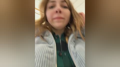 Media: A blurred, close-up video of a young woman with fair skin and light brown hair, wearing a green plaid shirt under a striped cardigan. Background is a dimly lit indoor space.