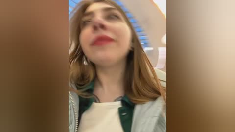 Media: Video of a young woman with fair skin and shoulder-length brown hair, wearing a light green jacket and white shirt, looking up in an indoor setting with blurred background.