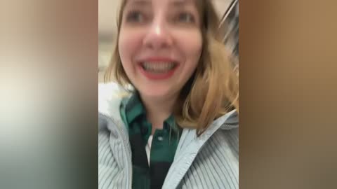 Media: Video of a smiling Caucasian woman with light skin, shoulder-length blonde hair, wearing a striped shirt and plaid tie, captured from a slightly blurred, close-up angle.