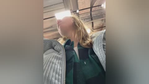 Media: Video of a blonde woman with a blurred face, wearing a green and gray plaid jacket, standing inside a rustic barn with wooden beams and natural light streaming in.