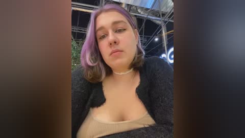 Media: Video of a young woman with light skin, shoulder-length pink hair, and a fair complexion. She wears a black cardigan over a beige top, with a visible cleavage. The background shows an indoor setting with industrial elements.