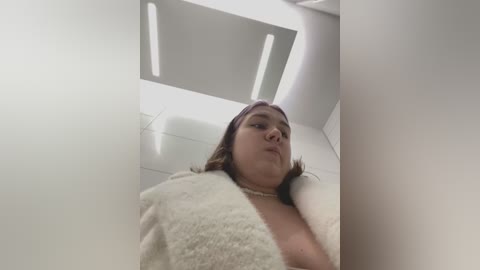 Media: A video captures a woman with fair skin, wearing a pink headband and a fluffy white bathrobe, standing in a modern, bright bathroom with white tiles and recessed lighting.