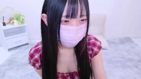 Media: Video of a young East Asian woman with long black hair, wearing a pink face mask, red plaid top, and sitting on a grey carpeted floor. Background features a white dresser and soft lighting.