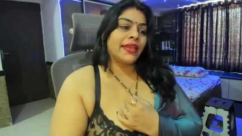 Media: Video of a South Asian woman with medium skin tone, wearing a black lace bra, sitting on a bed in a dimly lit room with floral curtains and a gray chair.