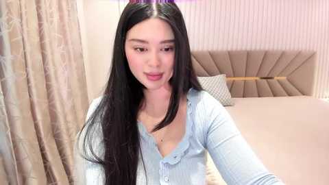 Media: Video of a young East Asian woman with long black hair, wearing a light blue button-up shirt, standing in a modern bedroom with beige curtains and a tufted headboard.