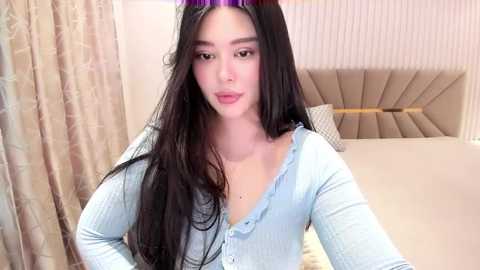 Media: Video of a young East Asian woman with long black hair, fair skin, wearing a light blue cardigan and a purple headband, posing in a modern, beige upholstered bedroom with a tufted headboard.