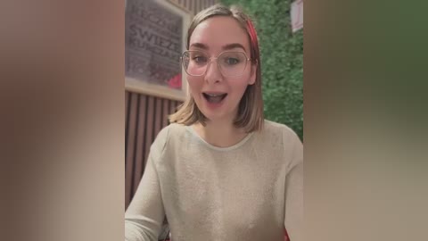 Video of a young woman with light skin and short blonde hair, wearing glasses and a beige sweater, looking surprised in a cozy, indoor setting with green foliage and wooden decor in the background.