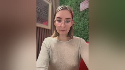 Video of a young, fair-skinned woman with light brown hair, wearing glasses and a beige sweater, sitting in a cozy, modern cafe with green wall art and wooden decor.