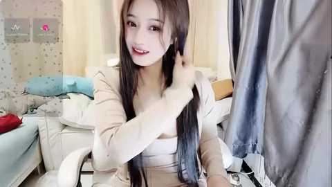 Media: Video of a young East Asian woman with long, straight black hair, sitting on a bed, holding a hairbrush. She wears a beige top and has a cheerful expression. The background shows a messy bedroom with white walls and a blue bedspread.