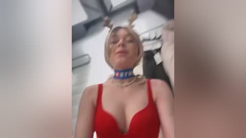 Video of a fair-skinned, blonde woman with large breasts wearing a red bra and a blue collar, standing in a dimly lit room with a blurred background.