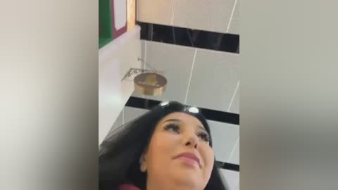 Media: Video of a woman with long black hair, wearing a red top, standing in a modern room with white and black tiled walls, a circular light fixture, and a framed green and red object on the left.