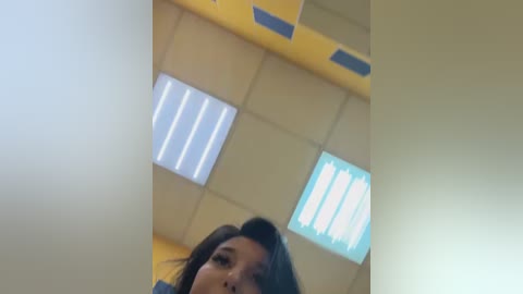 Media: Video of a young woman with straight, dark hair, captured from a low angle, standing in a brightly lit room with fluorescent ceiling lights and windows.