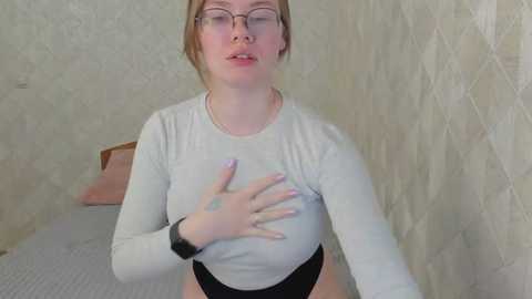 Media: Video of a fair-skinned, blonde woman with glasses, wearing a tight white long-sleeve top and black high-waisted panties, seated on a bed, touching her chest, in a beige-patterned room.