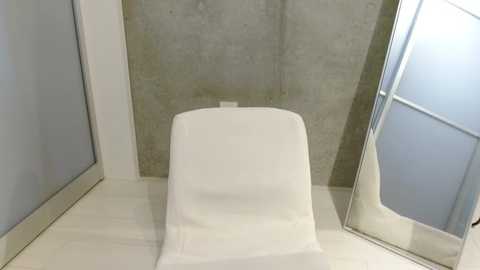 Media: Video of a minimalist bathroom with a sleek white chair against a concrete wall, a frosted glass shower door, and light-colored tile flooring.