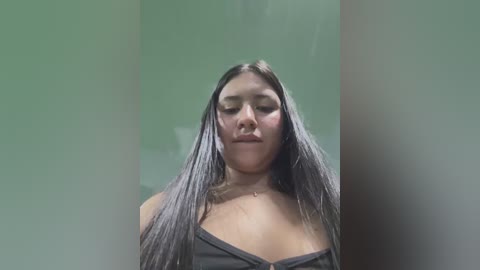 Media: Video of a young Latina woman with long, straight black hair, wearing a black strapless top, standing against a blurred, greenish-gray background.