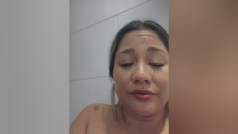 Video of a young Asian woman with light skin, brown hair, and closed eyes, possibly in a bathroom, wearing minimal makeup. She has a small tattoo on her upper arm.