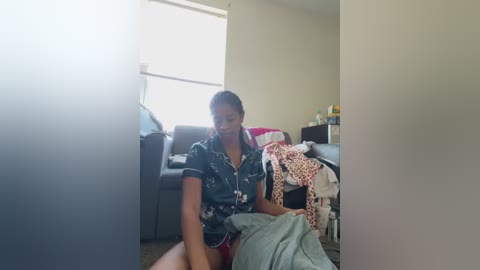 Media: Video of a young woman with medium-dark skin, dark hair, and a medium build, sitting on the floor in a messy living room. She wears a floral shirt and red shorts, holding a white towel. Background includes a gray couch, a window, and scattered clothes.