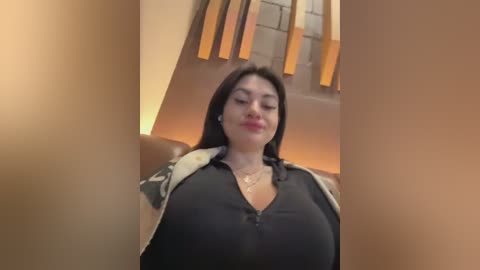 Media: Video of a curvy, light-skinned woman with long black hair, wearing a low-cut black top, sitting in a dimly lit room with beige walls and wooden blinds.