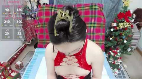 Media: A video of a woman with black hair in a bun, wearing a red, cut-out top, sitting in front of a festive Christmas tree. Background includes a plaid chair, wrapped gifts, and a \"Merry Christmas\" sign.