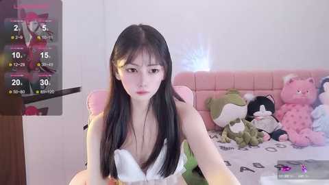 Media: Video of an East Asian woman with long black hair, wearing a white dress, sitting on a bed with stuffed toys, playing a video game.