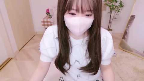 Media: Video of a young Asian woman with long brown hair and a white mask, wearing a white top with a cartoon print. She's indoors, standing in a hallway with beige walls, a potted plant, and a checkered chair in the background.