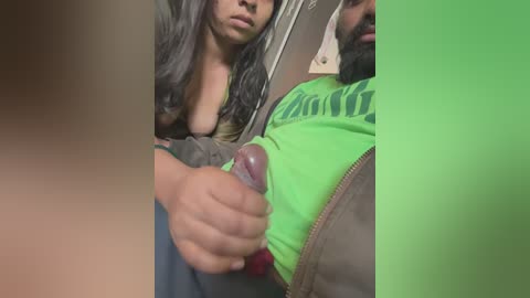 Media: Video of a woman with long black hair and a green shirt, holding a man's erect penis, partially visible. Background shows a blurred, neutral setting.