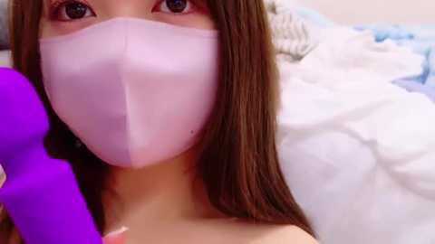 Media: Video of an Asian woman with long brown hair, wearing a pink face mask, holding a purple vibrator. Background shows a white and blue bedspread.