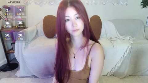 Media: A video of an East Asian woman with long, straight red hair, fair skin, and a slender physique, wearing a tan tank top. She sits on a beige couch in a softly lit room.