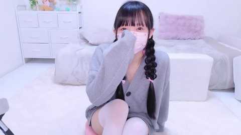 Media: A young East Asian woman with long black braids, wearing a pink face mask and a grey cardigan, sits on a white floor in a minimalist room with a white bed and dresser.