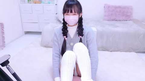 Media: Video of an Asian woman with long black braided pigtails, wearing a light grey blazer, white turtleneck, white mask, white thigh-high socks, seated on a light carpeted floor in a minimalist, pastel-colored room with a bed and white dresser.