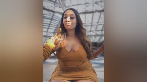 Media: Video of a voluptuous Black woman with long, wavy brown hair, wearing a low-cut, mustard-yellow dress, posing confidently with one hand on her hip. Background shows an industrial, arched ceiling.