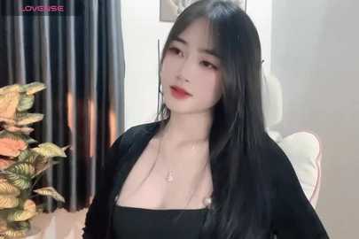 Media: Video of an East Asian woman with long black hair, fair skin, and red lipstick, wearing a black top, sitting indoors with a potted plant and curtains in the background.