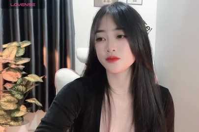 Media: Video of an East Asian woman with long black hair, fair skin, and red lipstick, wearing a black cardigan, sitting in front of a floral arrangement, white curtain, and white wall in a modern indoor setting.