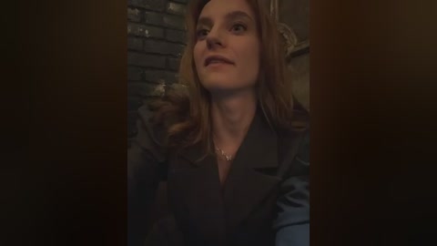 Media: Video of a young woman with long, wavy brown hair, wearing a dark blazer, standing in a dimly lit, brick-walled room, with a neutral expression.