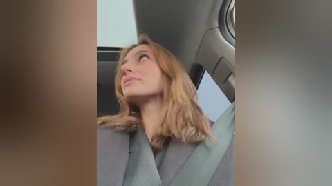 Media: Video of a young blonde woman with shoulder-length hair, wearing a gray jacket, sitting in a car. She looks upward, eyes closed, with a serene expression. The car's interior is visible in the background.